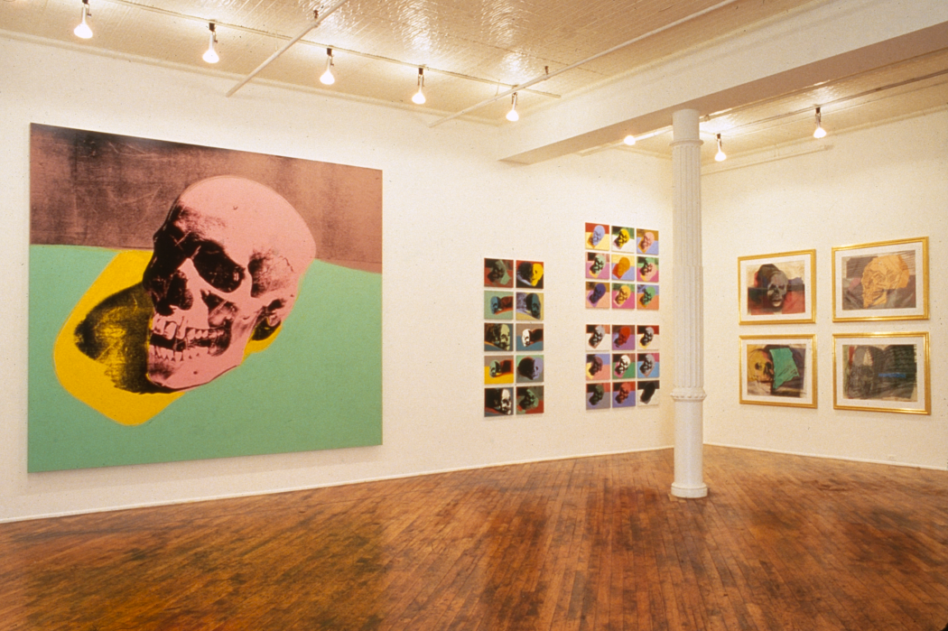 A large painting of a skull in aqua, yellow and pink fills most of a white wall. To its right are grids of smaller colorful skull images.