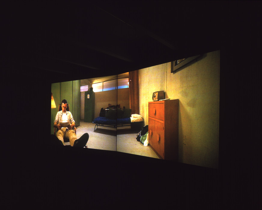 A two-channel projection featuring a man reading a newspaper in a room with small, high windows and a bed, chest of drawers, and radio.