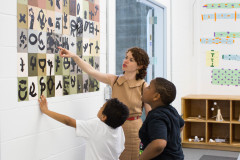 A Dia educator points to a square in a paper quilt of black icons and symbols on the wall as two youth attentively gaze upwards.