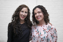 Two women smile for a photo against against a white brick backdrop, both have curly brown hair and one wears a black blouse and the other wears a pinks floral patterned blouse.