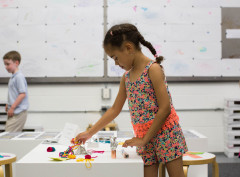 A young girl wearing a pink floral romper stands in a classroom and looks down at the art materials on her desk and holds a piece of coppr and string.