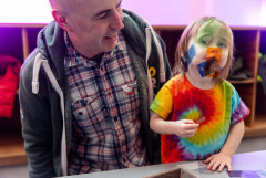 An adult looks, smiling, at a young child who, wearing a tye-dye shirt, looks at the camera while sticking small transparently colored rectangles to their face.