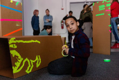 A kneeling child in a room full of other adults and children uses yellow tape and scissors to decorate a cardboard box with a large-mouthed animal.