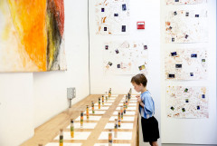 A child peers over a long table with two rows of tall thin glass vials containing various colors of seeds or beans. Works on paper are mounted on the far wall and ad large yellow, red and black canvas hangs on the wall adjacent to the table.