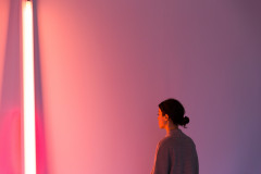 A neon work leans vertically in a corner and casts a pink and purple glow across the wall and a woman's face who stands nearby.
