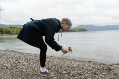 A woman bends over, magnifying glass and plant in hand, by a body of water to examine something on the rocky shore.