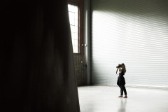 A dark shape diagonally bisects the image and reveals a photographer with long hair dressed in black who is small in scale comparison to the naturally lit warehouse space surrounding her as she takes a picture.