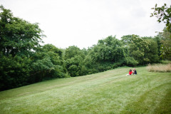 Two women sit in the distance on a grass with trees surrounding the grassy area.