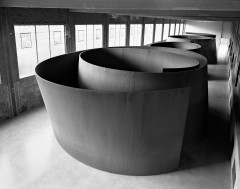 Large square windows are evenly spaced along one wall of an industrial room where a row of four large, curved sculptures consume the visible space in this black-and-white image.