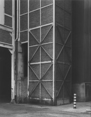 Black-and-white photograph of a corner of a brick building's exterior featuring a lattice of vertical, horizontal, and diagonal metal beams affixed to the brick.