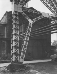 Black-and-white photograph of a brick building behind a massive set of intersecting vertical and diagonal metal trusses and four metal pipes suspended from the building in mid-air.