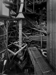 Black-and-white photograph of a factory setting with numerous sets of pipes, support beams, and staircases.