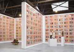 Paper works installed in a locked grid on a wall, each with a bright red background with two tan pages of a notebook, each with uniquely collaged images, texts, pages, postcards, or handwritten notes. The works are installed on tall walls, organized in three-sized rooms, two of which are visible here. There are also sculptures of people and stuffed animals arranged on both the floor and pedestals in the center of the rooms.