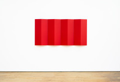 A red metal rectangular sculpture mounted on a white wall. The sculpture is a red metal sheet that is bent and folded into a symmetrical zig zag, installed so that the form alternates between extending into the gallery and then receding to touch the wall, starting and ending on the wall.