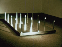 Stainless steel spikes in a five by three grid protrude upward from a stainless steel base on the floor of a gallery space.