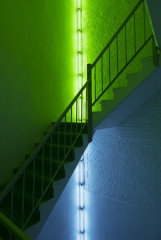 Two fluorescent artworks in corners on two floors of a stairwell, the lower piece is blue and the upper piece is green.