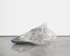 A folded, cone-shaped, iridescent, glass object rests on a reflective, cement surface in front of a white background.