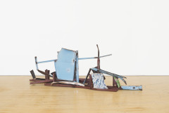 A horizontally oriented, long sculpture made of blue, red, and multicolored metal parts rests on a wooden floor.