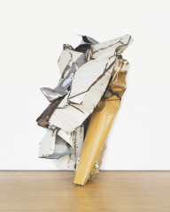 A crumpled-and-folded, white-and-yellow, metallic object rests on a wooden floor in front of a white background.