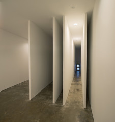 Two television monitors display camera output at the far end of one of five narrow, hallway-like spaces of varying width, formed by thin white walls.