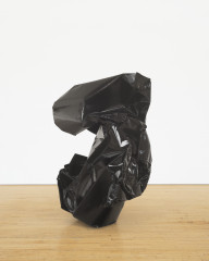 A crumpled sculpture made of black metal rests on a wooden floor.