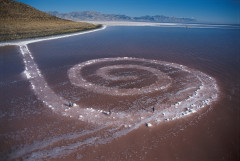 An aerial view of a large spiral made of rocks that runs counterclockwise from the shore into a pink lake.
