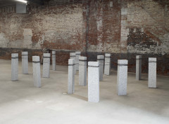 Fifteen upright, rectangular blocks of speckled granite are bound together with steel wire. The sculpture sits on a concrete floor in front of a brick wall.