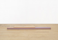 A thin, rectangular peach beam is stacked on a longer, pink beam on a wooden floor in front of a white wall.