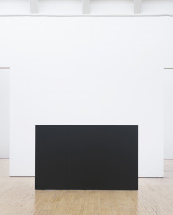A wall-like, rectangular, dark sculpture with two vertical seams is centered on a wooden floor in front of a square white wall.