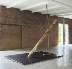 A long wooden log hangs from a thick rope above a large rectangular steel plate with small rocks sandwiched between the plate and concrete floor.