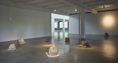 Seven large rocks rest on beige pillows and are arranged in a loose circle on a concrete floor with a single lightbulb hanging above and a spotlight to the right.