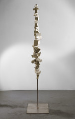 Balanced on a bronze pole and attached to a flat base is a thin vertical sculpture made of several wooden blocks of varying sizes that are stacked on top of one another.