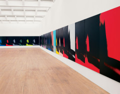 Twenty-five canvases hang edge to edge and low to the floor along a wall and in the corner of a gallery. Each painting features an identical abstract mark painted in black, gray, red, green, yellow, pink, purple, or blue. 