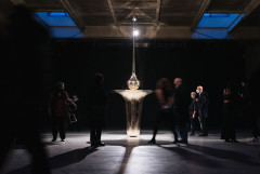 A half dozen people stand in a dark room around a shiny brass sculpture lit by a spotlight. The bottom form resembles the end of a horn, pointing up, the top form, resumbling a mute or a clapper, hangs from above.