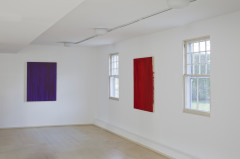 Two paintings, a purple rectangular canvas and a red rectangular canvas and a on adjacent white walls between two windows.
