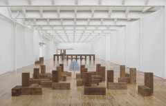 Many wooden columns and several gray bricks are positioned in different configurations such as archways and Tetris-like shapes across the wooden floor of a long white room.