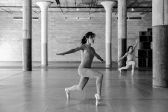 Black-and-white photograph of two dancers in leotards lunging to the right while en pointe with arms exended outwards. The dancer in the center foreground has arms pointed perpendicular to their body. The dancer in the right background has their left arm pointing diagonally upwards and forwards, and right arm pointing diagonally backwards and downwards.