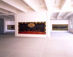 In the forefront a large rectangular painting hangs horizontally on a white wall. In the background, two large rectangular paintings hang on a white wall in the back of the same room.