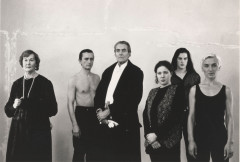 Black-and-white photograph of six people standing posed next to each other against a plaster wall in different states of dress, all wearing black clothing. The clothing ranges from white tie formal to a black tank top.