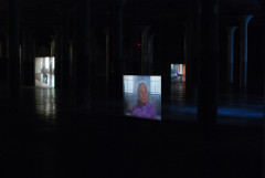 Three video projections of the same old woman from different angles are differerntly positioned throughout in a dark basement space.