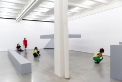 Four people dressed in different colored clothes occupy a white room with different sized grey rectangular prisms. The space is bisected by a white column and pipe. Two people are against a wall: one is seated with legs extended and the other stands, both staring across the room. One person sits adjacent to a hanging grey platform and gazes up at it. One person squats to look at another grey rectangular prism.