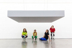 Four people in different colored clothes gaze up at a hanging grey platform in a white room.