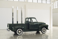 A vintage dark green Chevrolet pickup truck sits in profile in an open gallery space with a row of large windows in the background. Placed vertically in the truck's flatbed are circular, square, and triangular polished-stainless-steel rods.