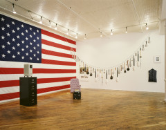 A room is filled with various artworks. The left wall is entirely covered by a large United States flag. In front of the flag is a black four-drawer metal filing cabinet with a portable white radiator on wheels placed on top. A lavendar sofa chair sits in the corner facing a CRT television set. From one side of the room to the other hangs a line of many different sized clubs.