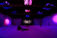 Several people sit, lie and stand in a large, dark room with purple and pink lighting. A projection of a multicolored abstract image is centered on the far wall, and sculptural objects hang on the left and right walls.
