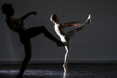 Two dancers in a darkly lit room, one in shadow in the foreground and the other illuminated in the back. Both are in the same position: standing on one foot en pointe in ballet shoes, with the other leg raised up, leaning back with both arms extended forward.