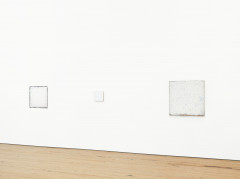 Three square white paintings hang on a white wall above a wood floor.