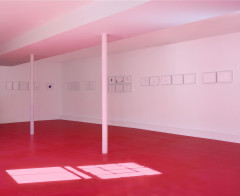 A white room is illuminated pink from natural light coming in from one window and reflecting off of the red floor. The window outline is visible on the floor. Multiple framed ballpoint drawings hang on the wall.