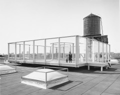 A black and white photograph of a woman and child in a roofless glass room on a platform installed on a rooftop, with a water tower just behind it, and the tops of square skylights surrounding it.