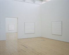 Two identically sized, square, white paintings hang on adjacent walls in a white room, and another square, white painting is seen on a white wall in the next room. Each painting has different sized horizontal stripes in alternating shades of white.